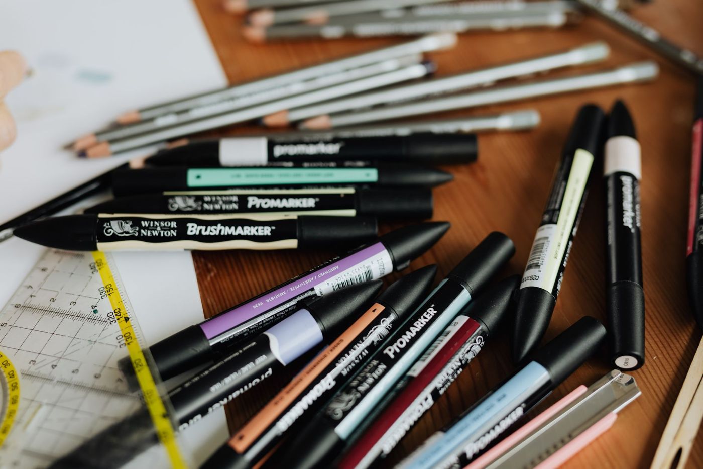 Dubbelzijdige Promarker-stiften in pastel en donkere tinten en potloden door elkaar op een houten tafel, naast een liniaal