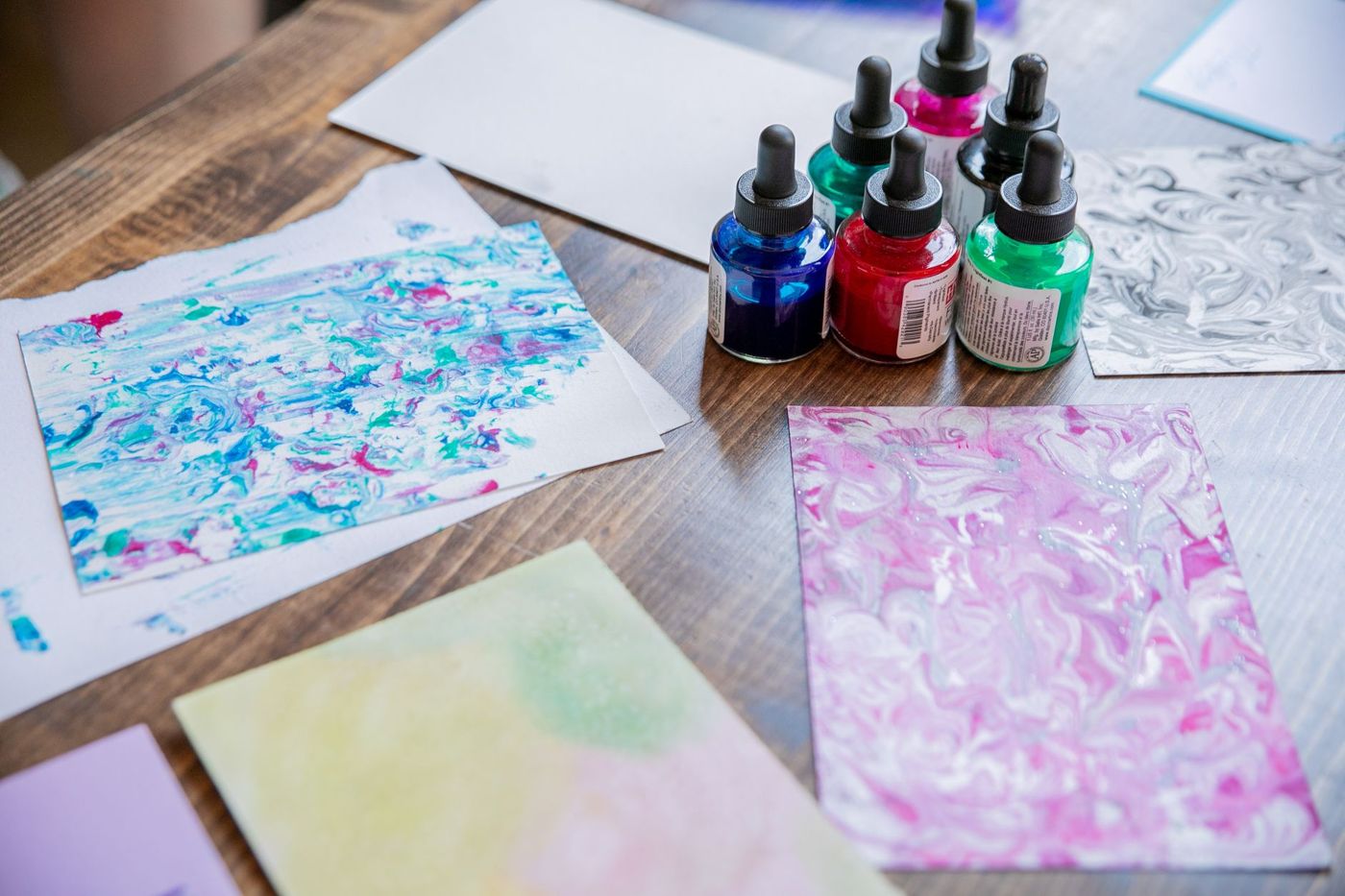 Zes druppelflesjes inkt in blauw, rood, groen en zwart naast gemarmerde papieren met roze en blauwe kleurverlopen