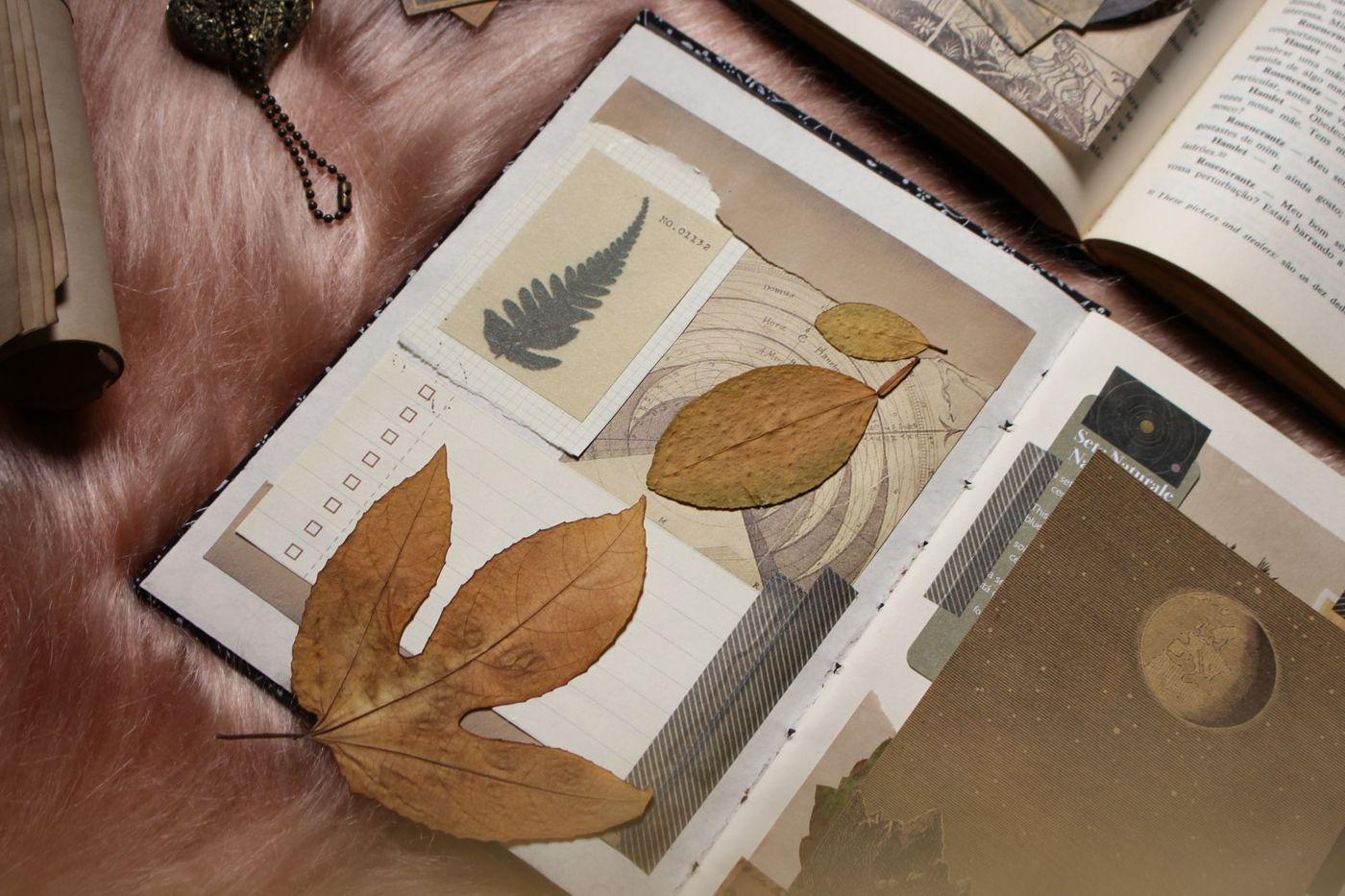 Opengeslagen junk journal met een collage van gedroogde bladeren, een varenmotief, oude kaarten en een maan, op roze vacht