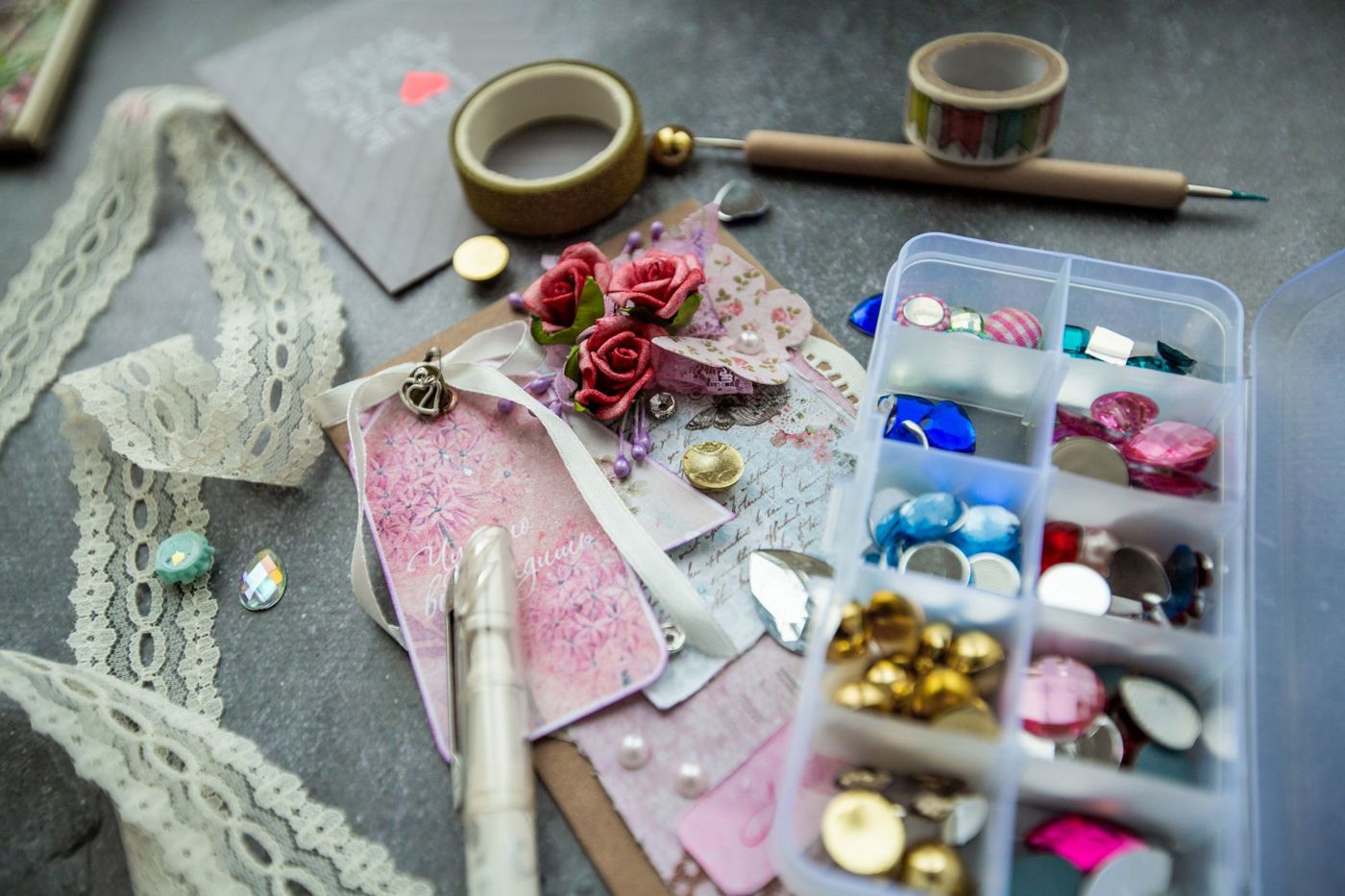 Papieren rozen op een roze label met lint, daarnaast kant, washi tape en een doosje met glinsterende steentjes en knoopjes