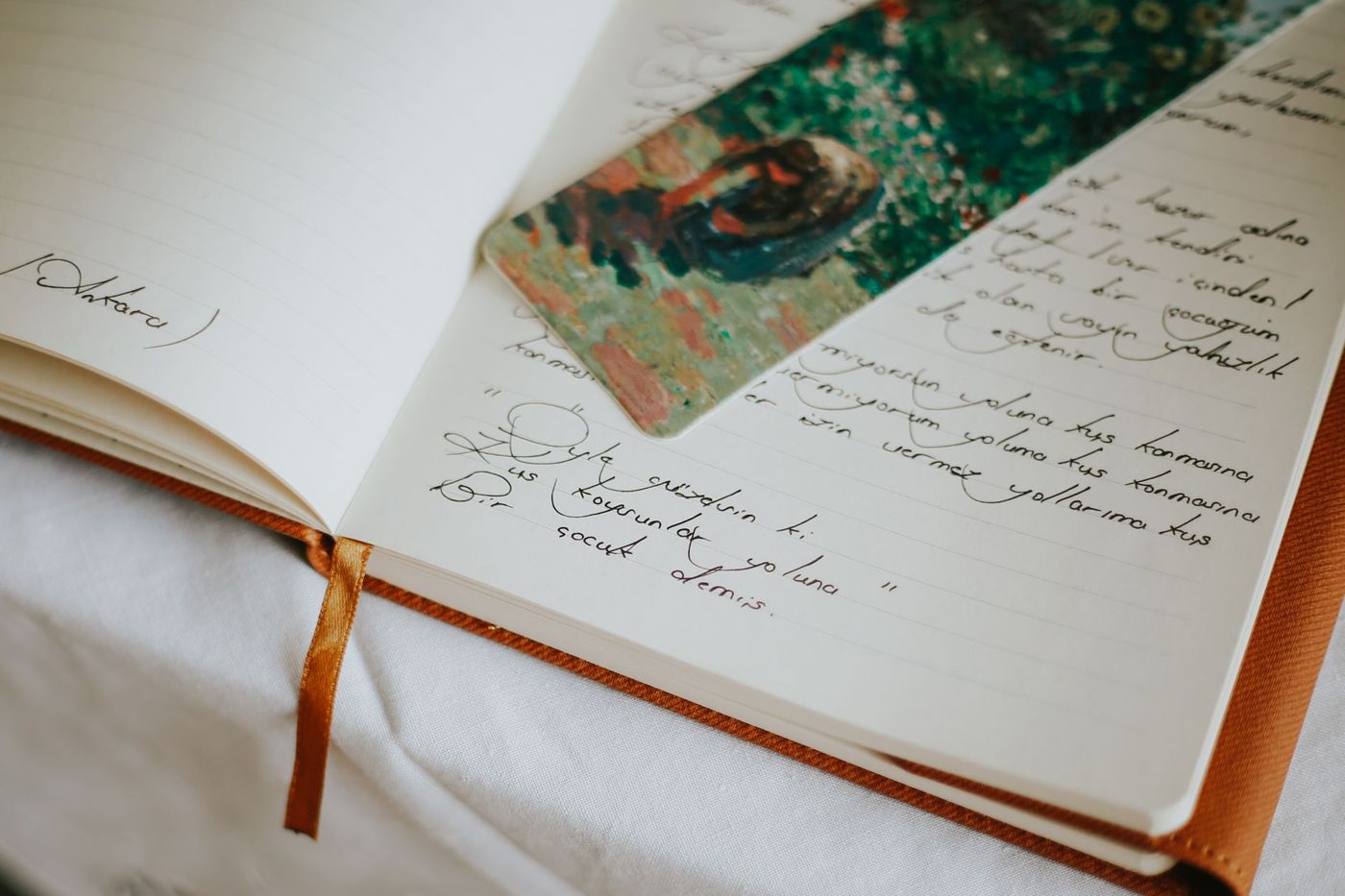 Opengeslagen schrift met handgeschreven tekst en een kaartje met een schilderij als bladwijzer, naast een oranje lint