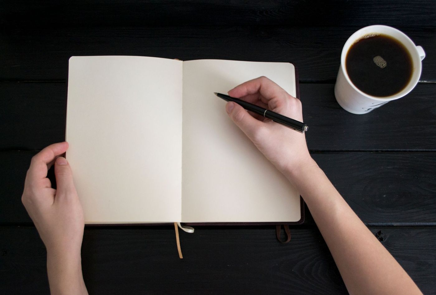 Twee handen boven een leeg opengeslagen notitieboek, met een pen en een kop koffie op een zwarte tafel