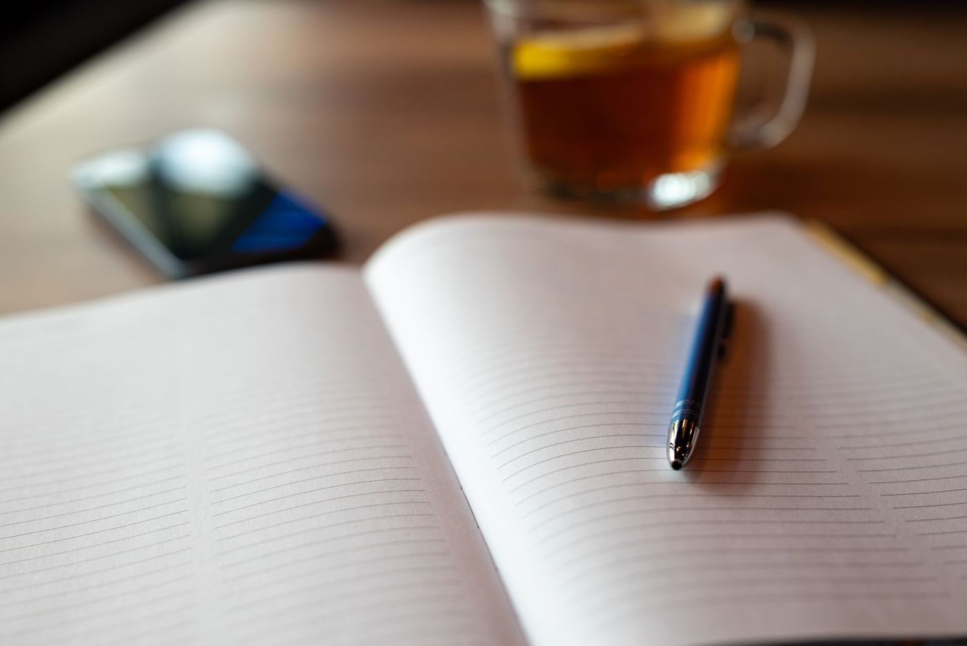 Opengeslagen gelinieerd notitieboek met een blauwe pen, onscherp een glas thee en een smartphone op een houten tafel