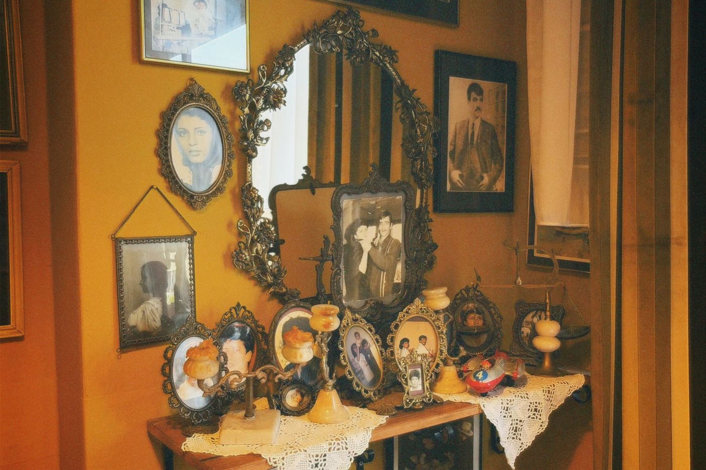 Tafel en wand vol oude familieportretten in sierlijke lijsten, met zwart-witfoto's van mannen en vrouwen en kantwerk eronder