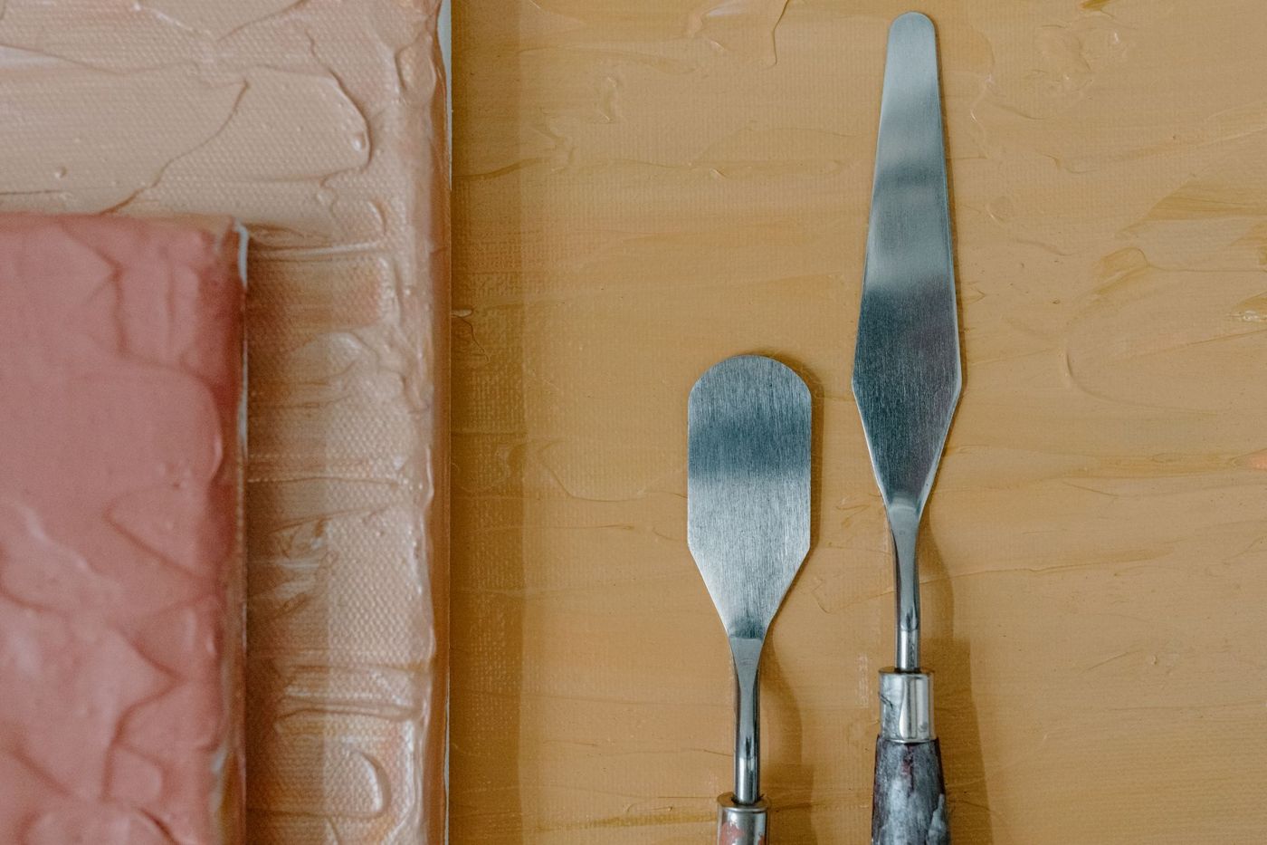 Twee paletmessen op een ondergrond met dikke verf in oranje en roze tinten, links een geschilderd canvas