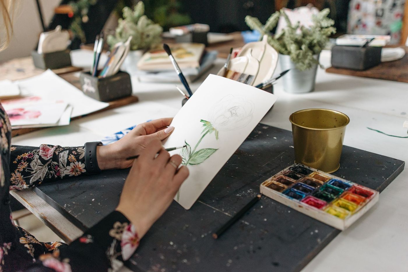 Twee handen schilderen met een penseel een groene bloemsteel op een vel papier, naast een waterverfdoos en potten penselen