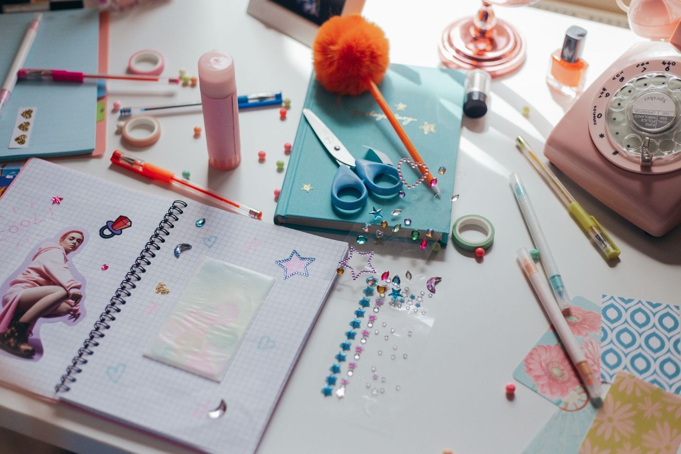 Witte werktafel vol scrapspullen: turquoise notitieboek met schaar en pen, stickers, washitape, lijmstift, roze telefoon