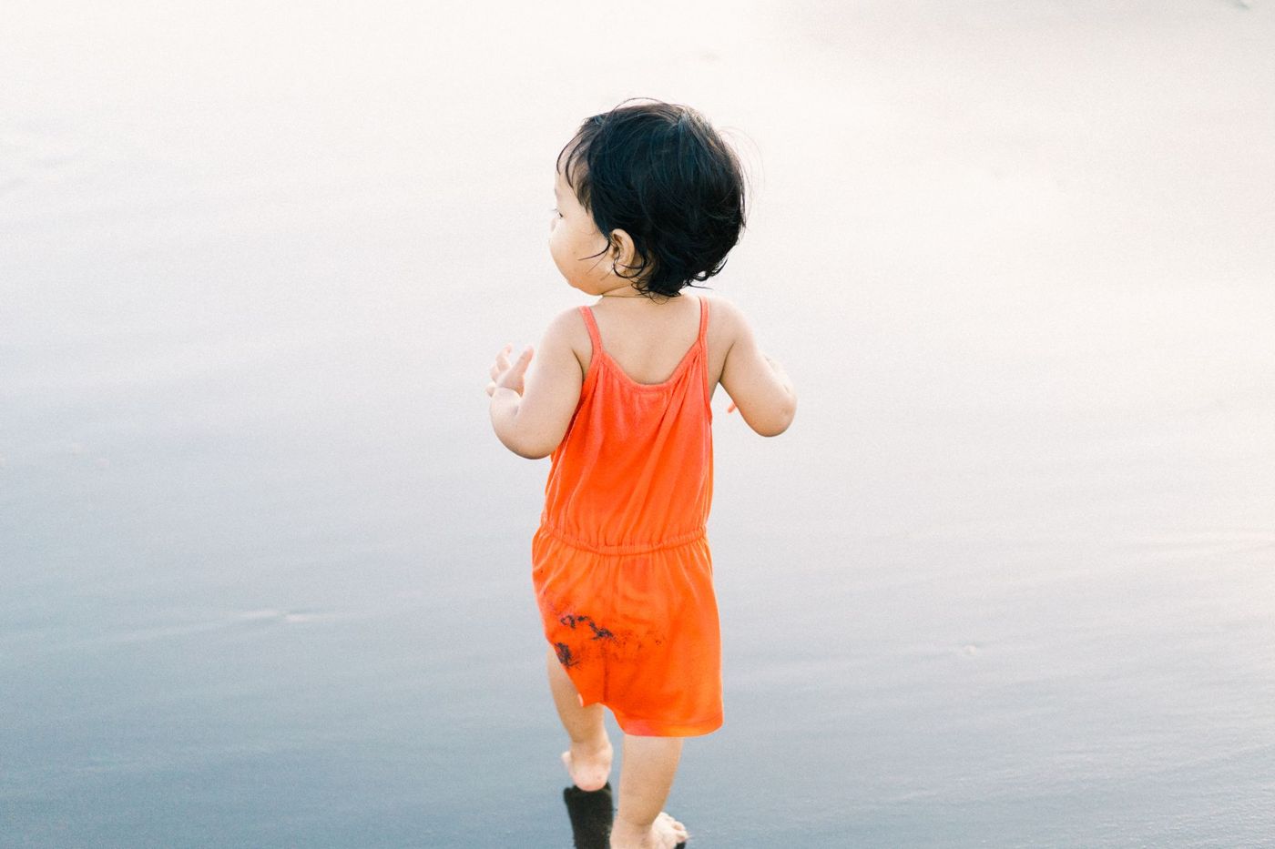 Peuter in een oranje jumpsuit loopt van achteren gezien over een rustig, glanzend strand