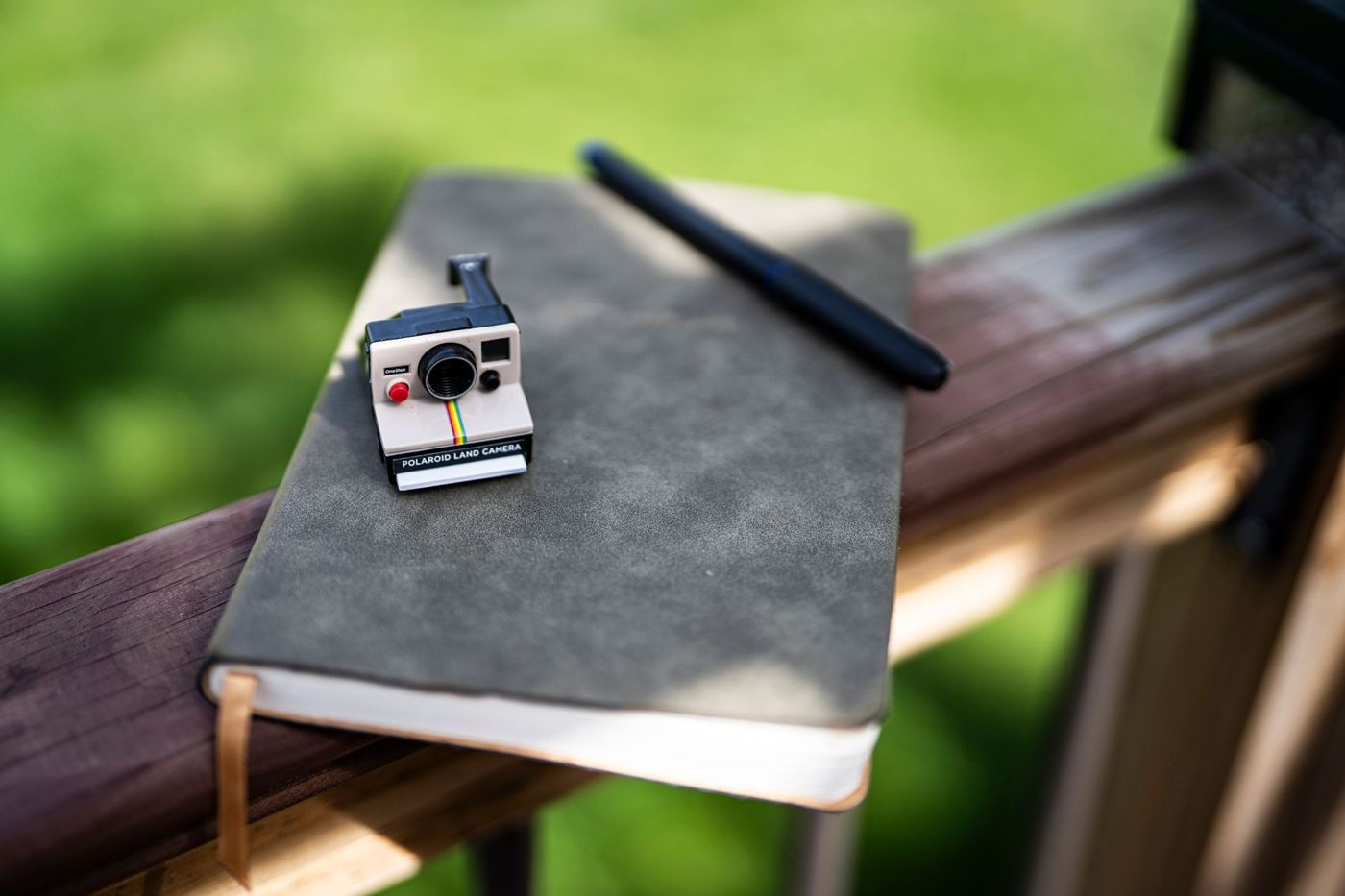 Klein model van een Polaroid-camera op een grijs notitieboek met een zwarte pen, op een houten reling in de zon