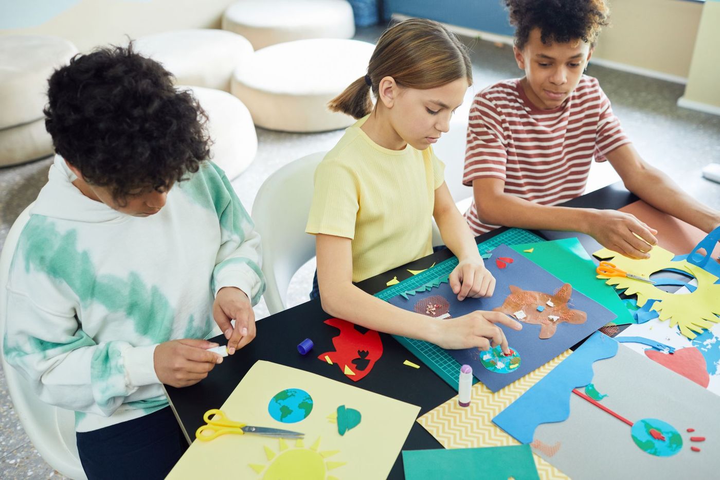 Drie kinderen plakken en knippen aan tafel met gekleurd papier, een schaar en een lijmstift