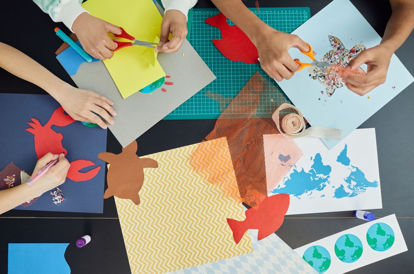 Kinderhanden knippen en tekenen aan tafel tussen gekleurd papier, een wereldkaart en een snijmat