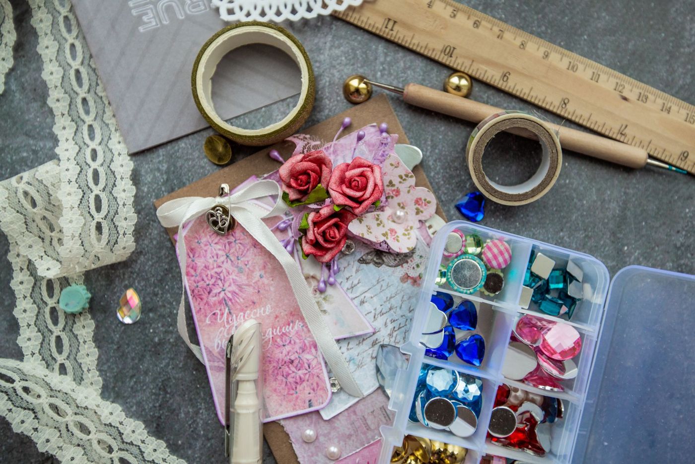 Scrapbookspullen op een grijze ondergrond: kant, washi tape, strass-steentjes in een doosje en een roze kaart met rozen