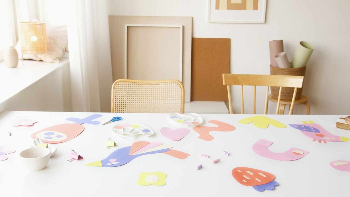 Witte tafel met pastelkleurige uitgesneden papieren vormen, een schaar en kleine knijpers bij een raam