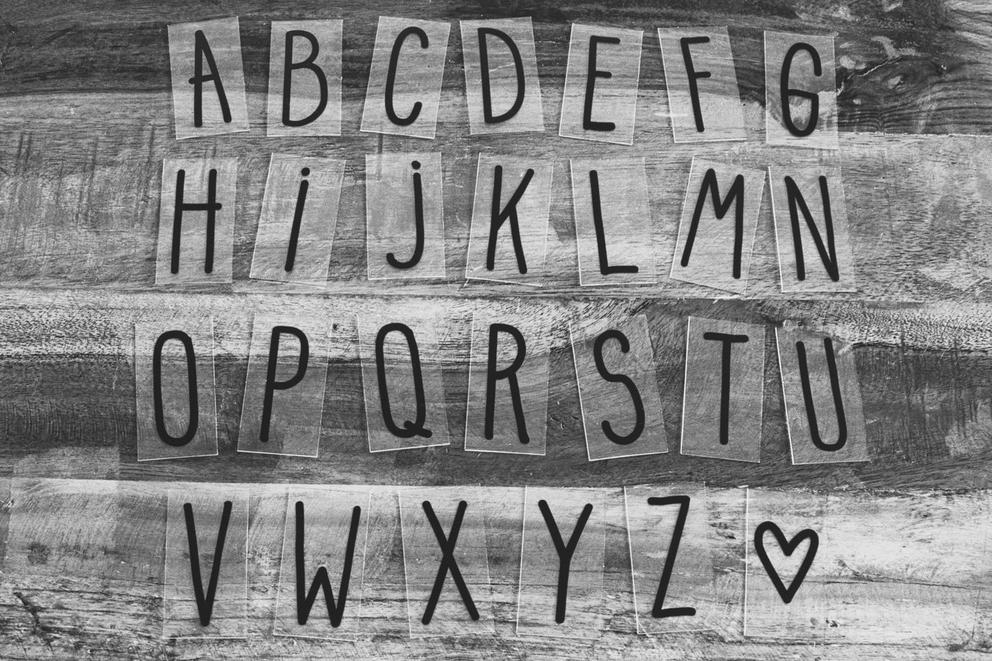 Zwart-witfoto van het alfabet in zwarte letters op doorzichtige strookjes op een houten ondergrond, met een hartje