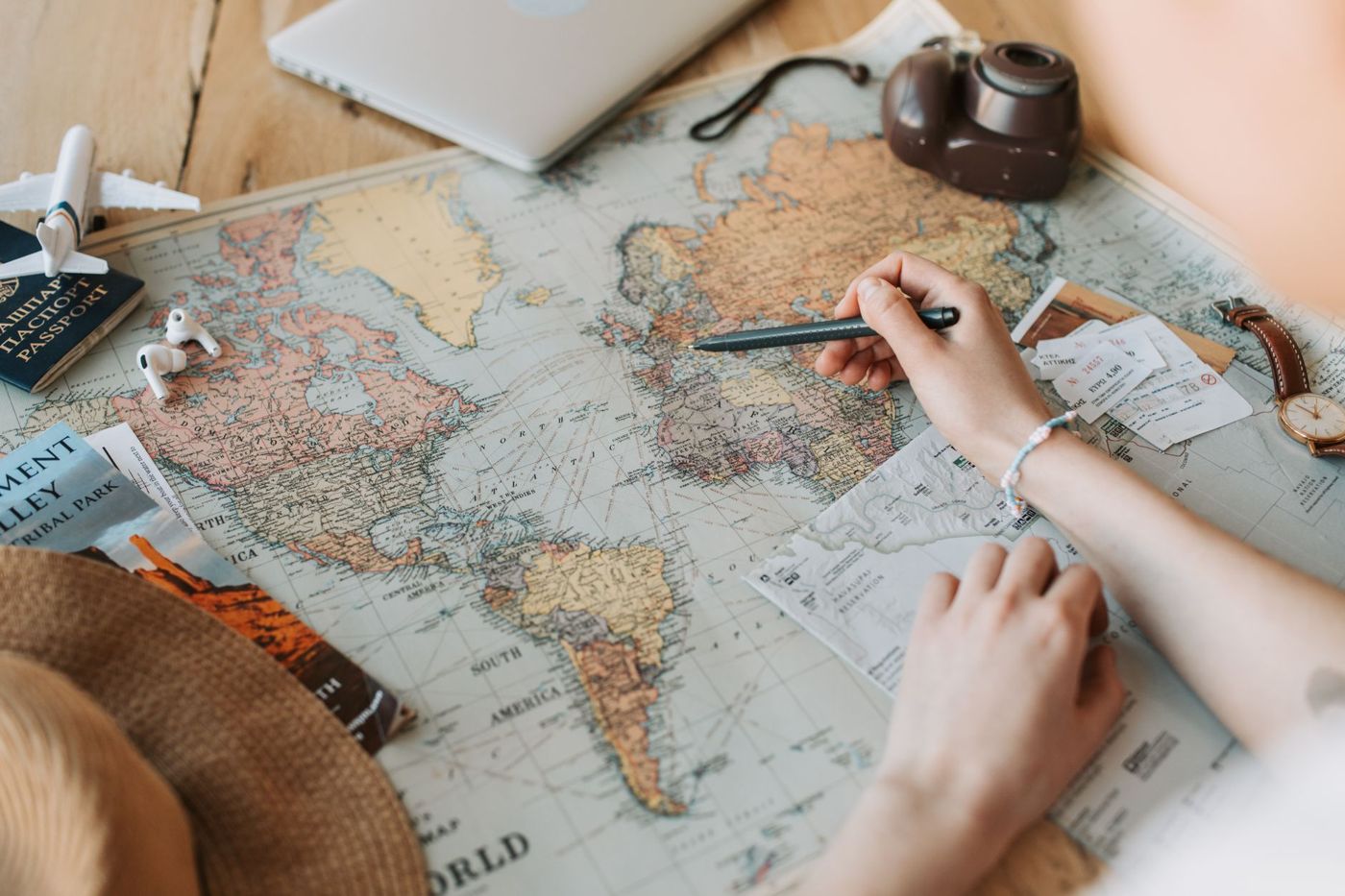 Hand wijst met een pen naar Europa op een wereldkaart, omringd door paspoort, camera, treinkaartjes, folder en strohoed