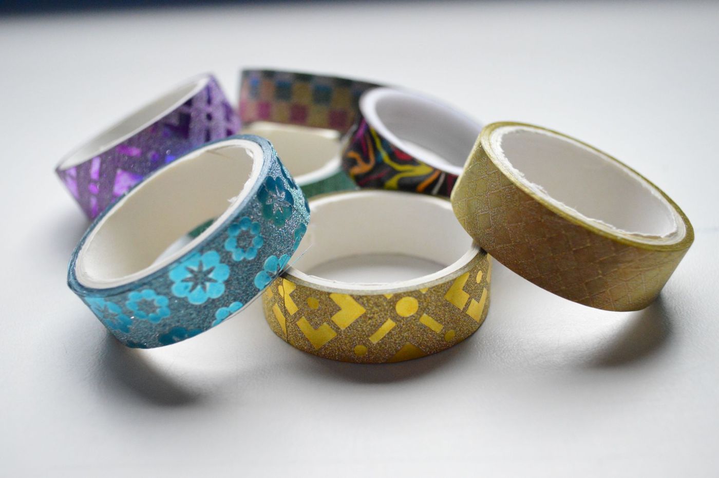 Zes rolletjes washi tape met glitter en patronen in blauw, goud en paars op een lichte ondergrond