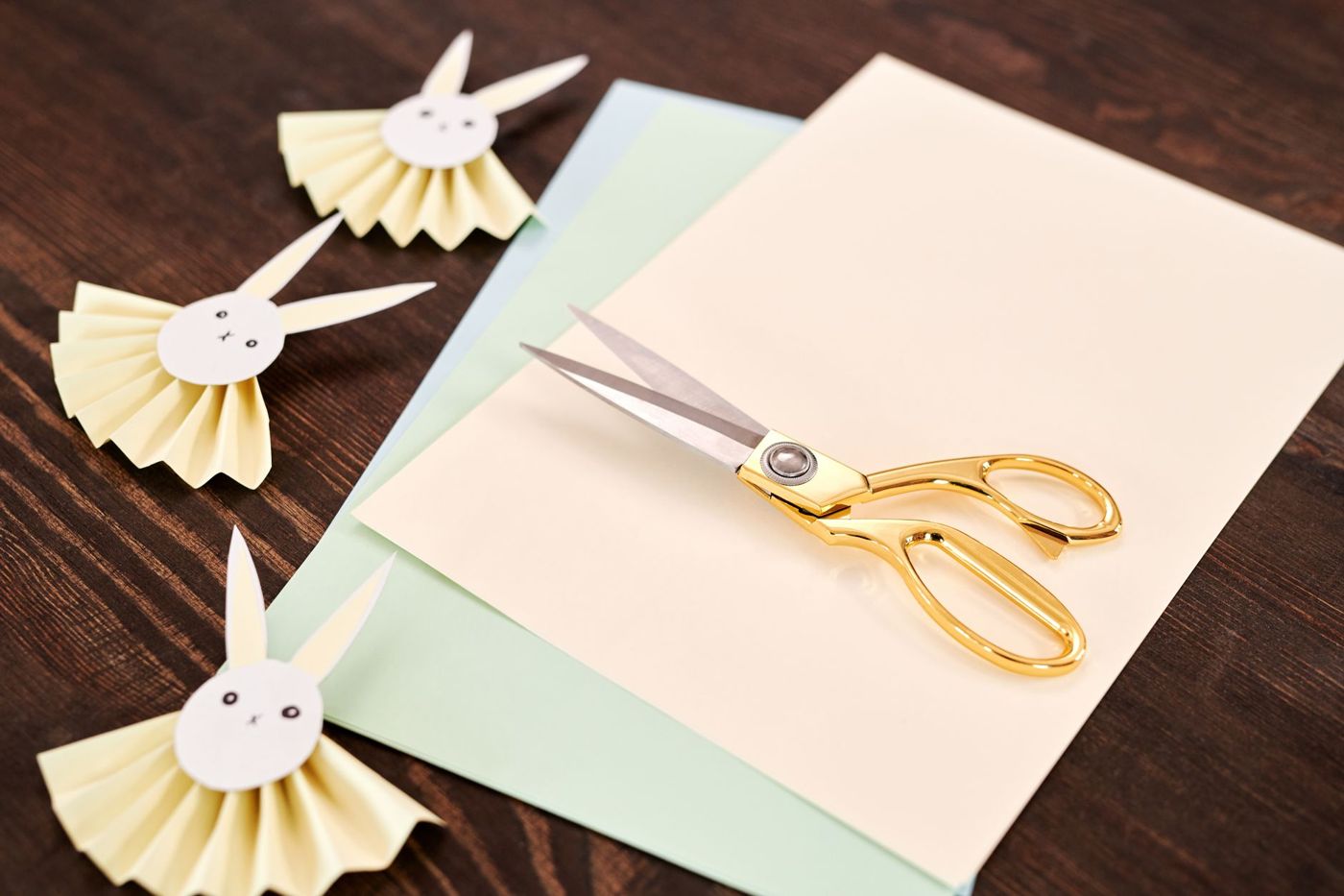 Gouden schaar op vellen crème, groen en blauw papier, naast drie papieren plooirozetten in de vorm van een konijntje