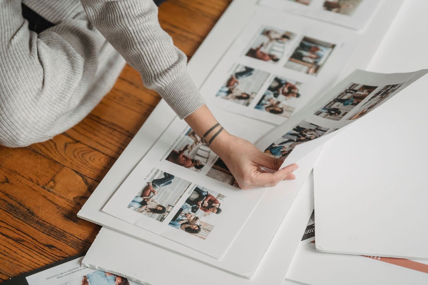 Een hand tilt een geprint vel met foto's op, boven andere vellen met fotolayouts op een houten vloer
