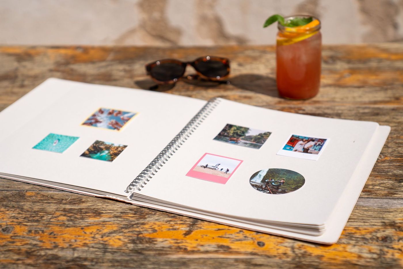 Opengeslagen spiraalalbum met kleine foto's op crèmekleurige pagina's, op een houten tafel naast een zonnebril en een glas