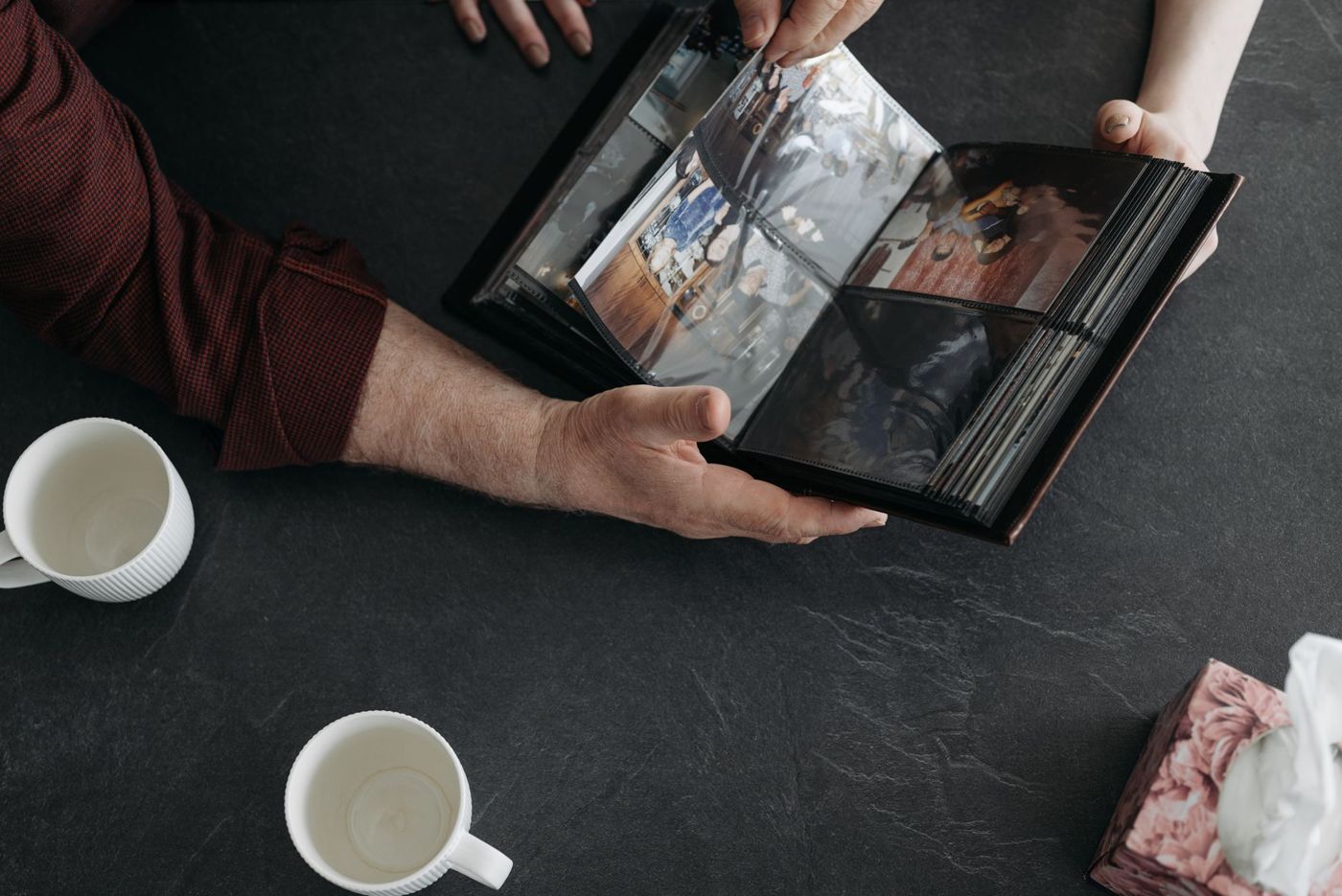 Handen bladeren door een fotoalbum met plastic insteekhoezen op een donkere tafel, naast twee witte mokken