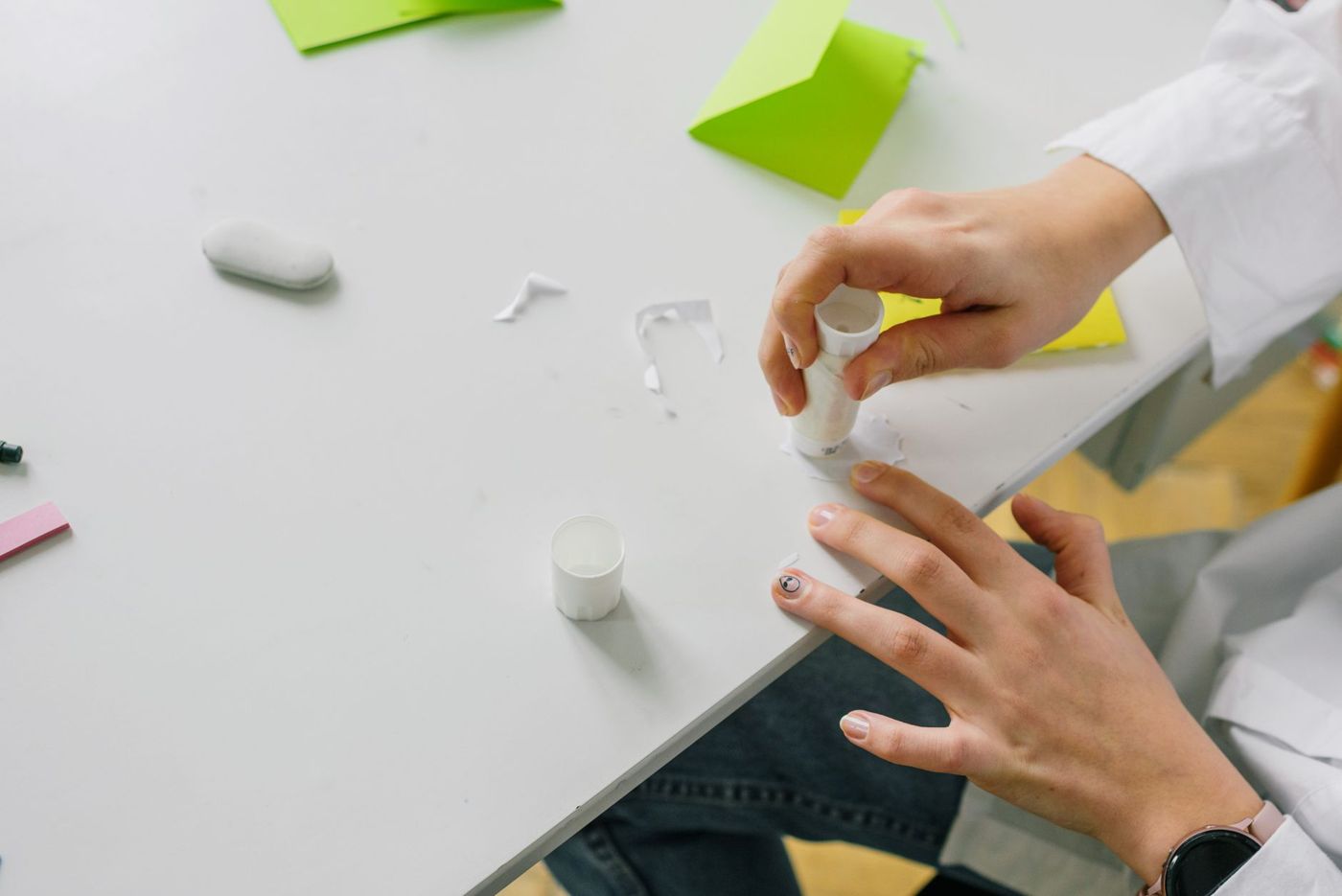 Twee handen houden een lijmstift vast boven een stukje papier op een witte tafel, met groene kaartjes en een gum