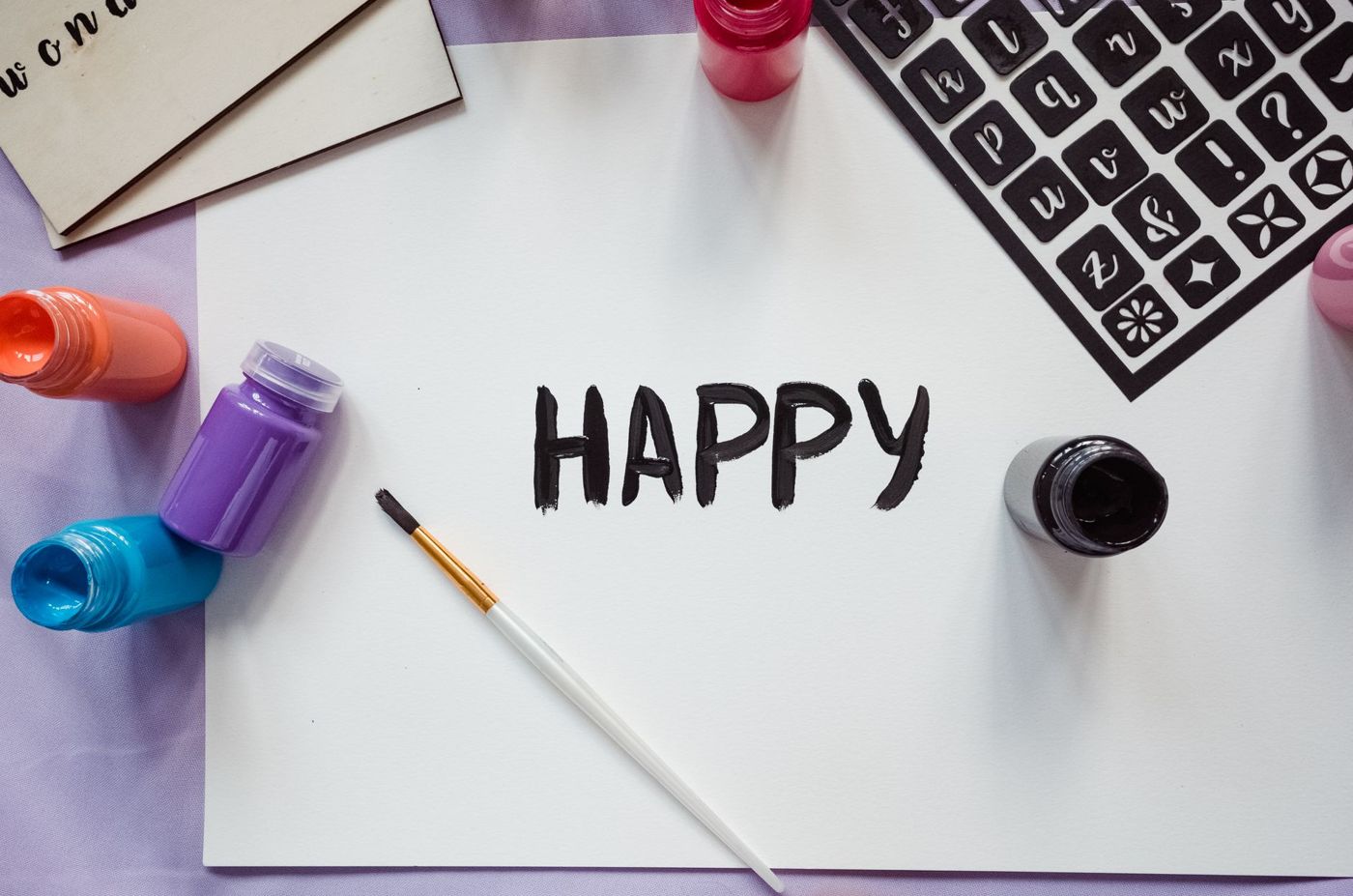 Het woord HAPPY in zwarte verf op wit papier, met penseel, potjes verf en een zwart stencilvel met letters ernaast