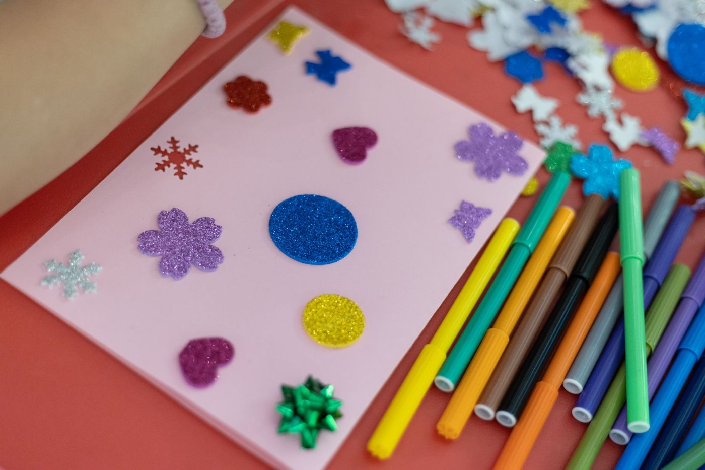 Roze kaartje met glitterstickers in hartjes, bloemetjes, sneeuwvlokken en cirkels, naast een waaier gekleurde stiften